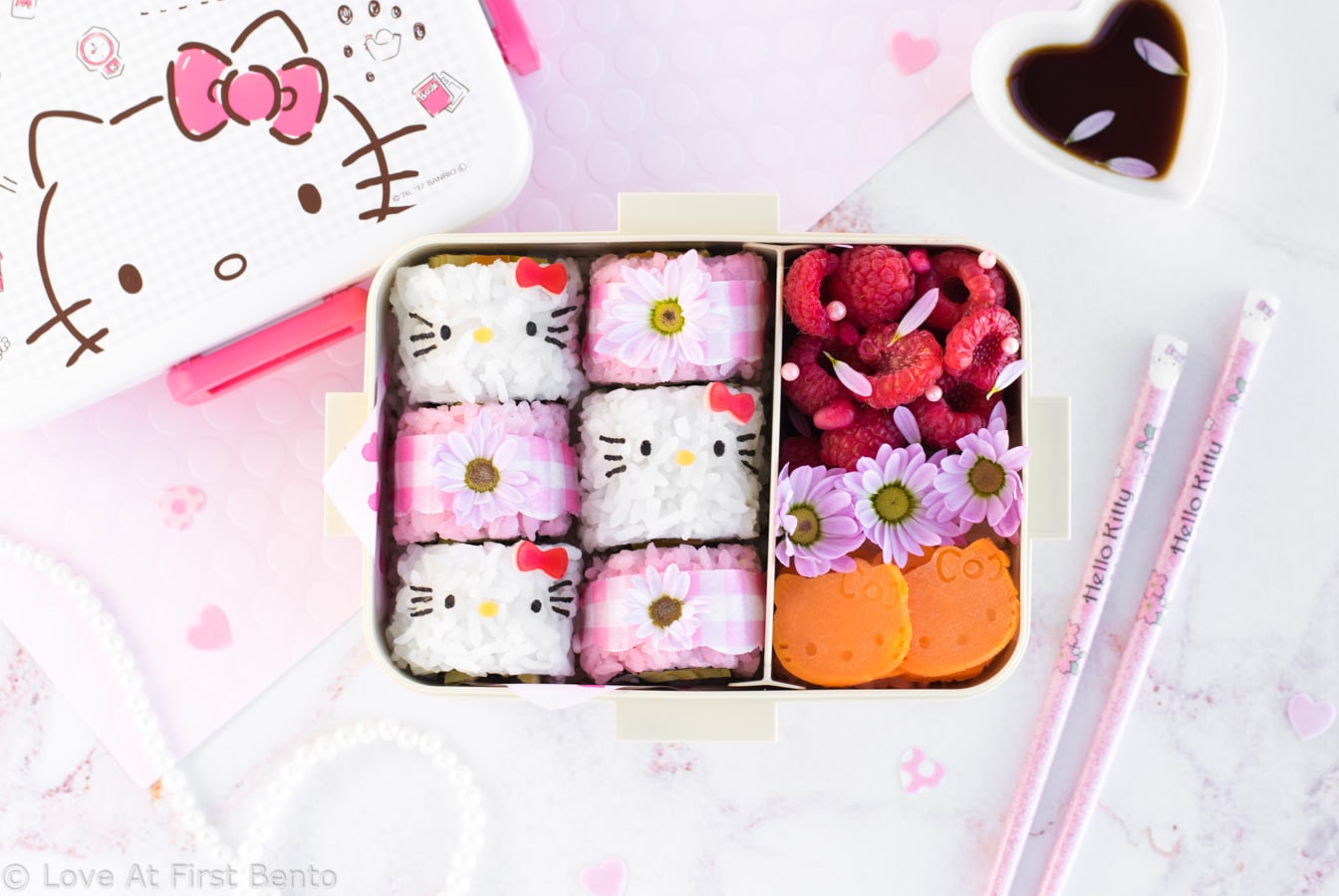 Hello Kitty Sushi Bento - Beautify your lunch with this Hello Kitty Sushi Bento! Filled with adorable Hello Kitty sushi rolls, as well as pink gingham flower sushi rolls, this pretty bento box is an edible dream come true for any Hello Kitty fan! Find out just how easy it is to make by visiting loveatfirstbento.com [character bento, kyaraben, deco sushi]